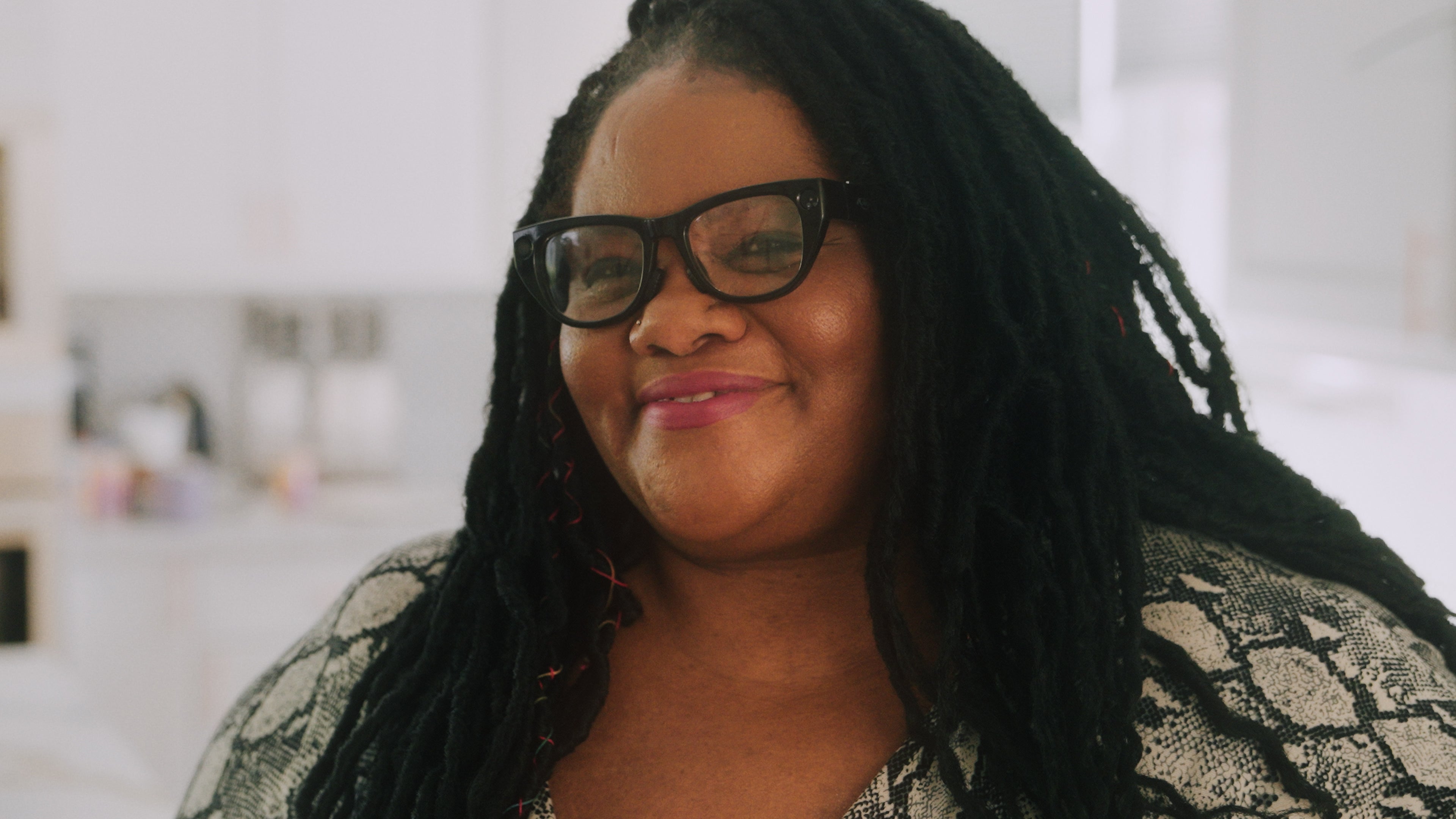 A smiling African American woman with braided hair wearing sleek EchoVision smart glasses in a blurred indoor setting. Her expression radiates confidence and independence, showcasing the stylish and natural fit of the device.