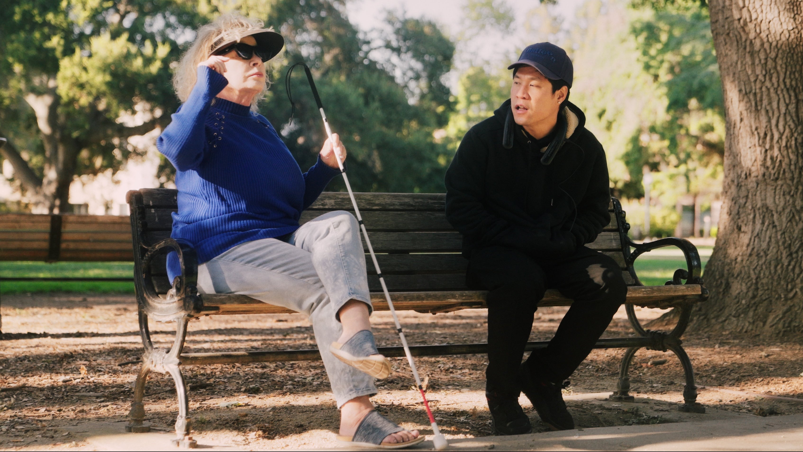A woman in a blue top and a man in black sit on a park bench surrounded by greenery. The woman, wearing EchoVision smart glasses, has her finger near the right temple after pressing the tactile button. The man is talking to her, illustrating how the device provides information without interrupting natural social conversation.
