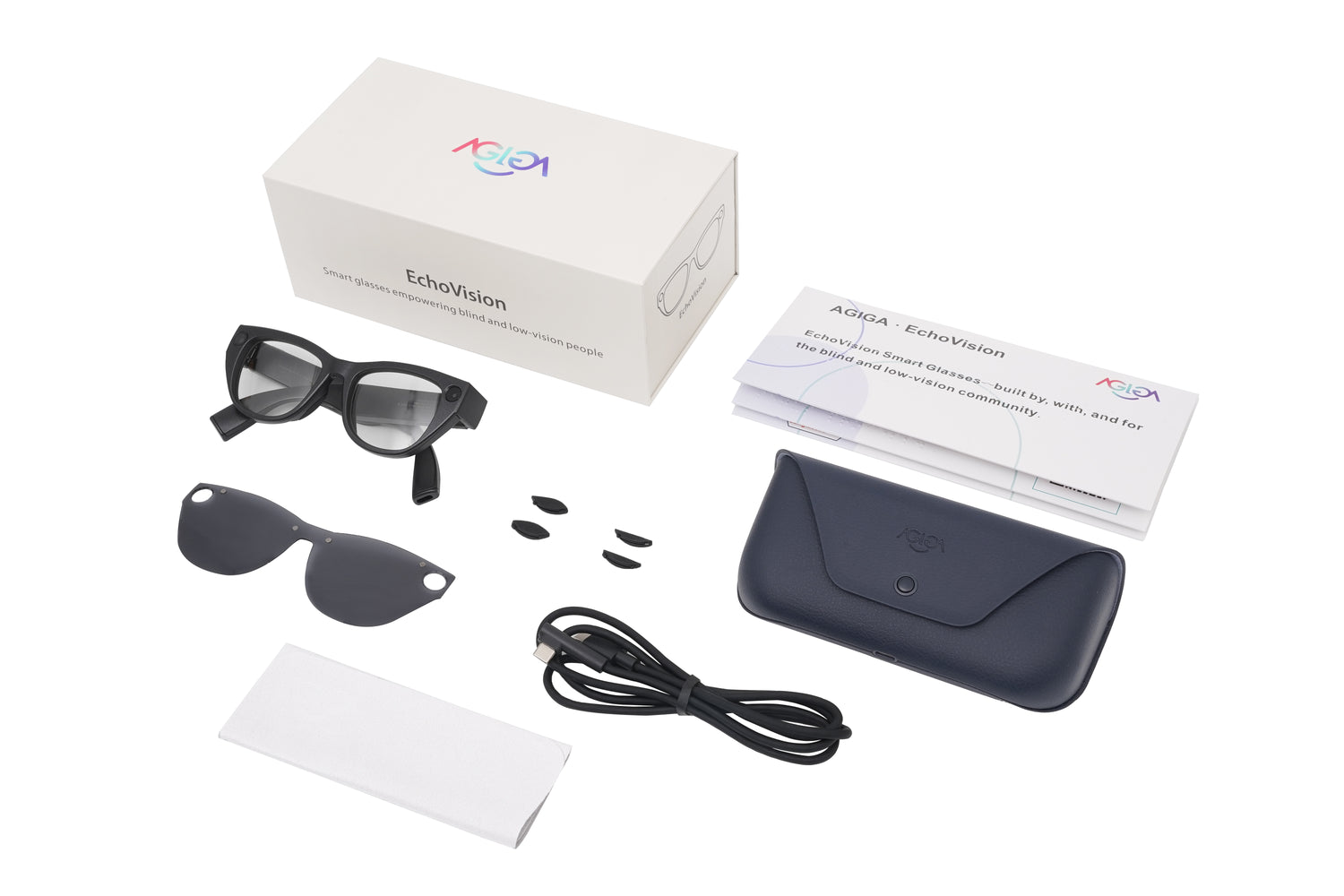 A top-down view of the AGIGA EchoVision packaging contents laid out on a white background. The display includes the AI-powered smart glasses, a sunglasses clip-on, a charging case, a USB-C cable, two pair of nosepads and a cleaning cloth. Pictured alongside the grey, branded carton box featuring the AGIGA logo is a 'Getting Started' guide with Braille.